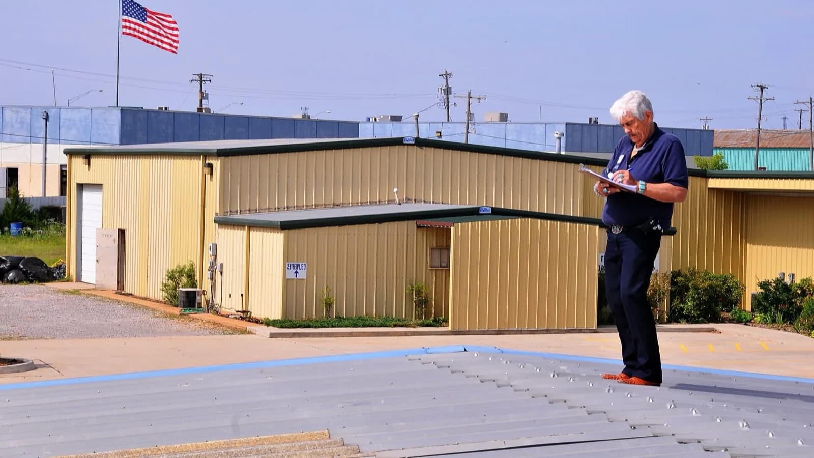 An inspector stands on a hail-dented metal roof, documenting storm damage on a clipboard.