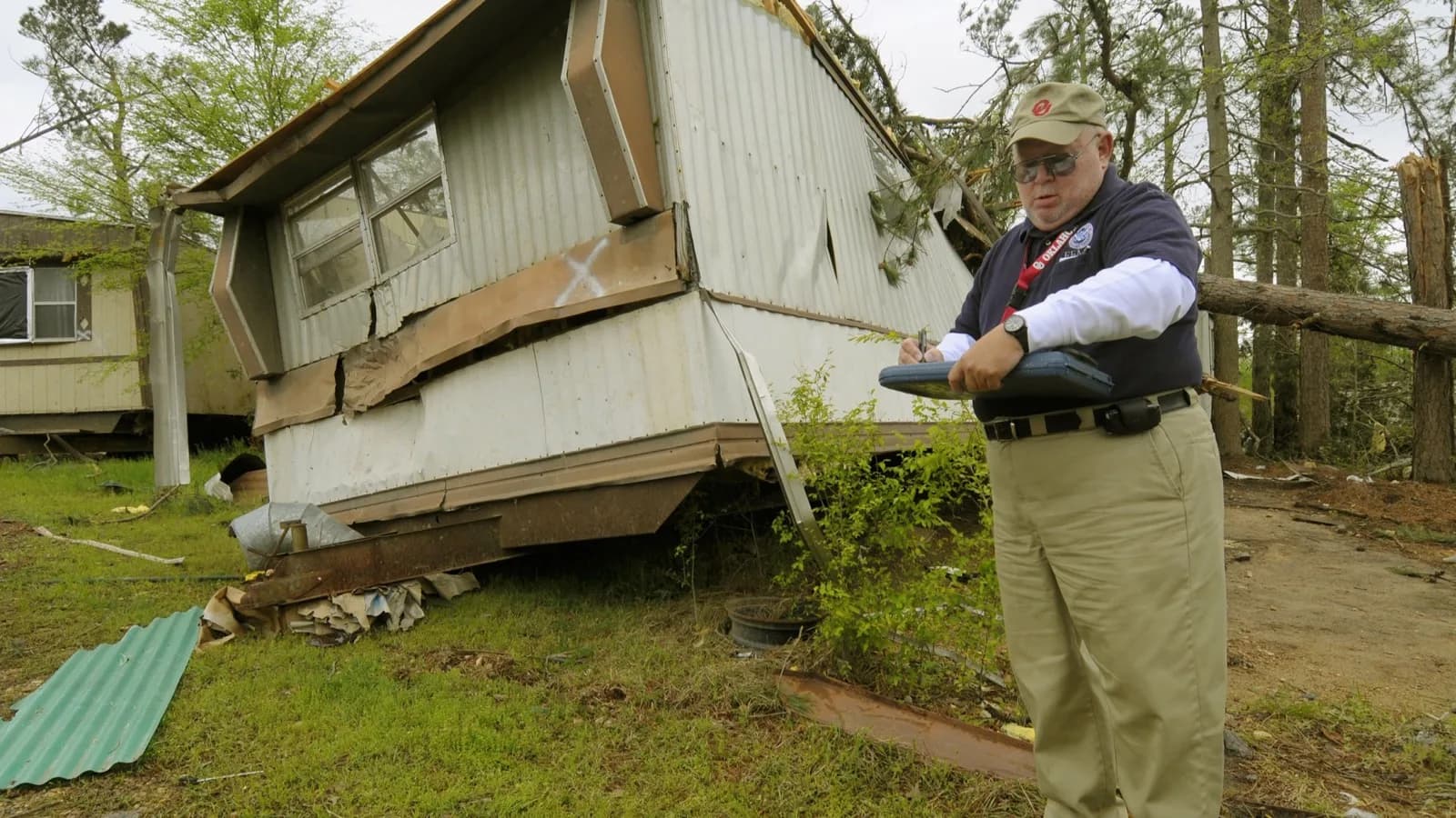 A FEMA damage-assessment officer stands beside a storm-wrecked mobile home, writing his findings on a clipboard.